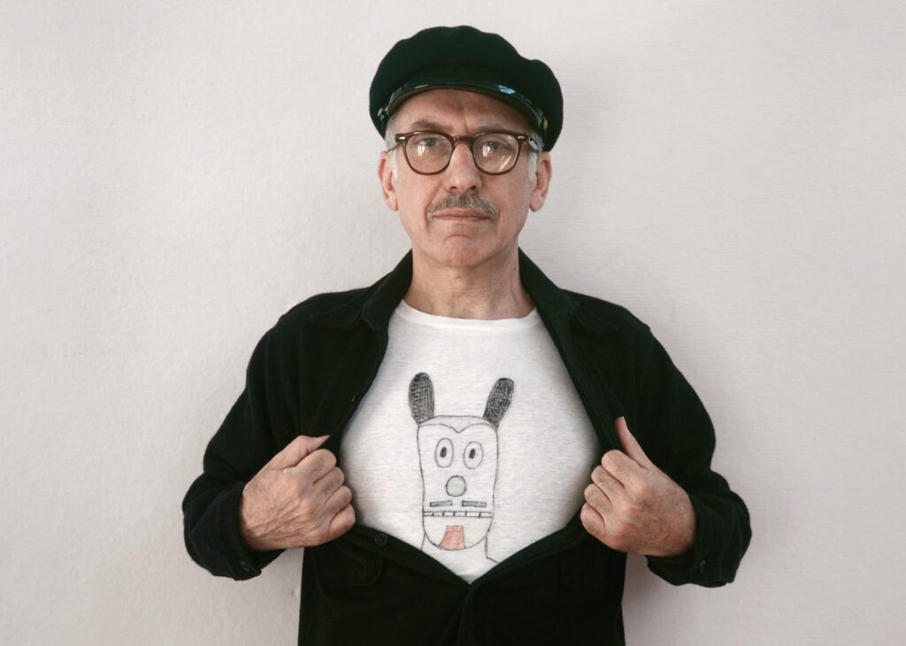 Photo of Saul Steinberg opening his jacket so you can see his t-shirt which has a hand drawn character on it
